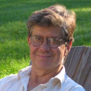 Photo of a middle aged white man with glasses sitting in a wooden chair out on a grassy lawn