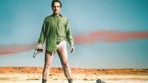 Walter from Breaking Bad, standing, facing the audience, in a green shirt and no pants, holding his gun.