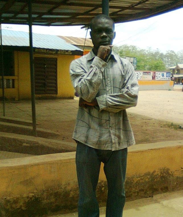 Chimezie Ihekuna (Mr. Ben) Young Black man in a tan collared shirt standing with his hand under his chin outside a building with eaves and a shade roof. 