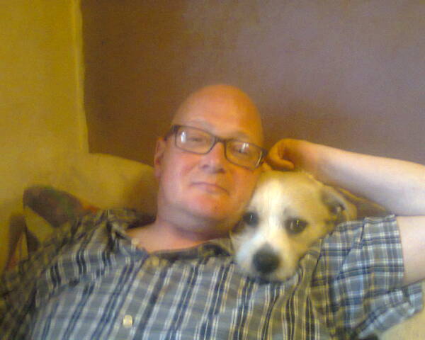 White man in a checkered buttoned top lying down with his arm up by his head, next to a dog.