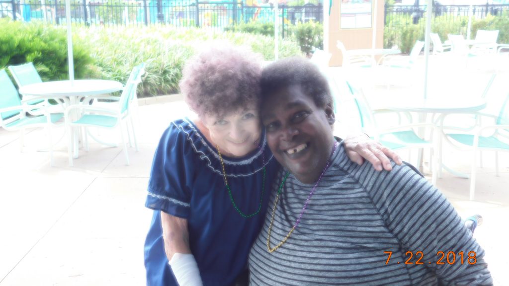 Middle aged Black man wearing a tee shirt hugging an older White woman, fellow contributor Joan Beebe, to his left. They're standing on concrete in front of some bushes and chairs near a swimming pool and both have necklaces of Mardi Gras beads.