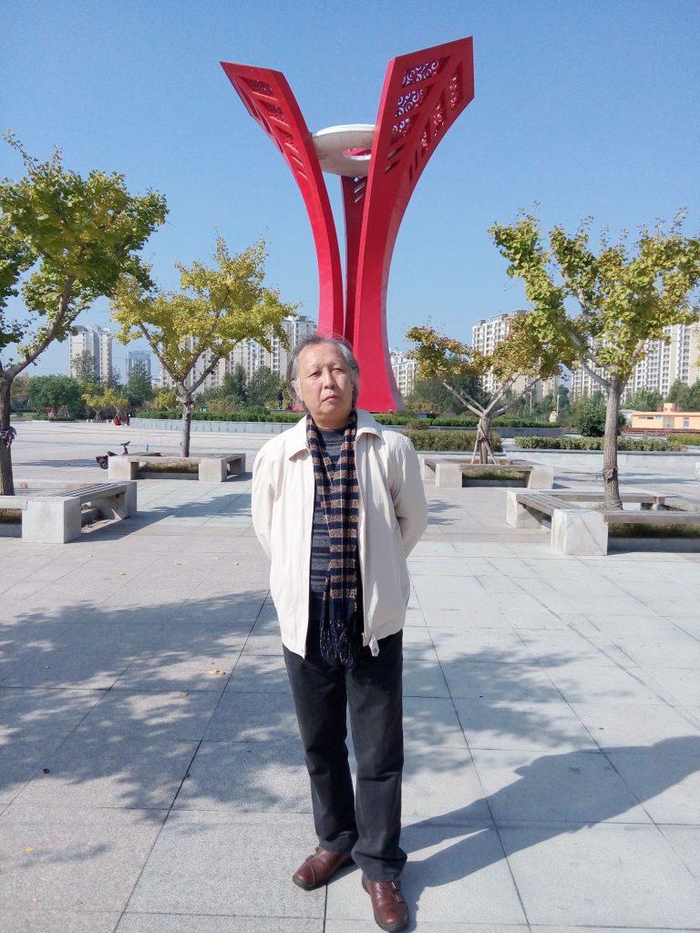 Middle aged Chinese man dressed in  slacks, brown shoes, a white coat and a scarf and striped shirt standing  in a city plaza, concrete with trees in planters. 
