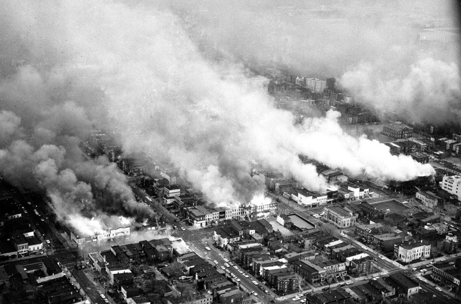 Black and white photo of smoke rising from burning buildings in Washington D.C. Aerial view over several city blocks. 