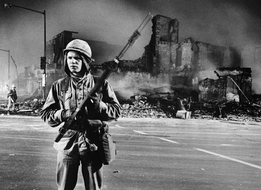Here, a trooper stands guard in the street as another (left) patrols a completely demolished building. White man with a large gun and a helmet stands on a street corner in front of a pile of rubble.
