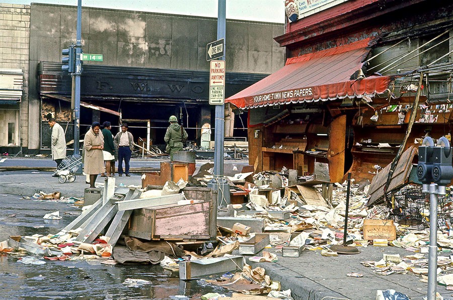 A few Black people with coats peruse a destroyed newsstand. Paper and wooden crates are strewn everywhere on the sidewalk and street in front of the storefront. Water pools in the street.