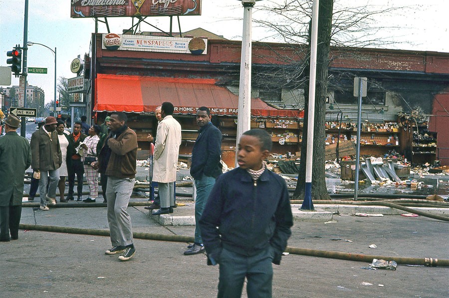 Young Black boy with a coat and jeans looks off to the side in another shot of the destroyed newsstand. More people are present, Black adults in coats and jeans. 
