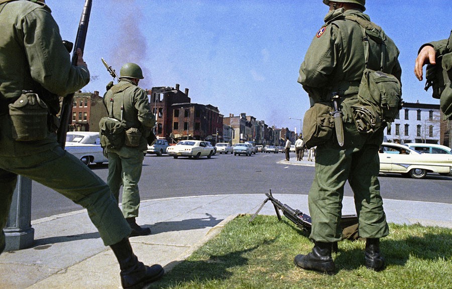 Green-uniformed National Guard troops stand guard with long guns on a street corner, facing some burned-out buildings. 