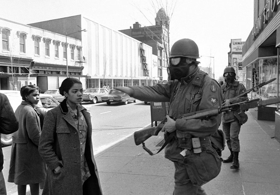 An armed, helmeted, uniformed National Guard officer waves away a young Black woman in a long coat as she looks back towards an area she's not permitted to enter. 