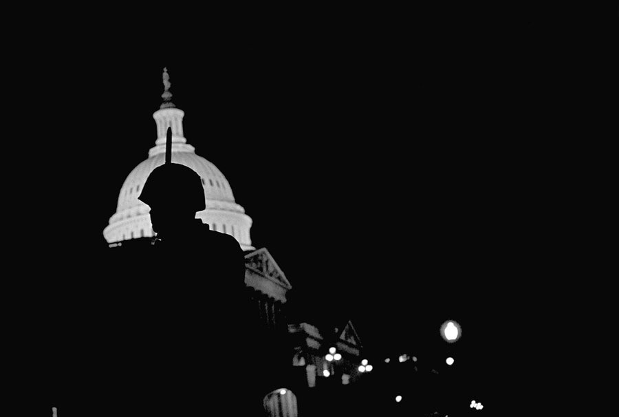 Silhouette of a National Guard officer standing up with his gun in his hands in front of the white lit-up Capitol building at night. 