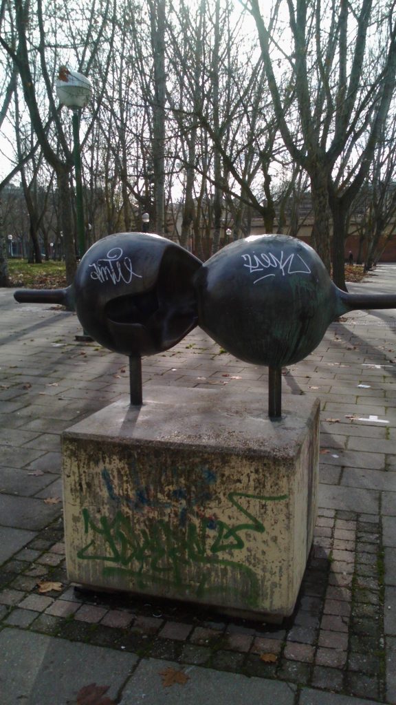 Metal sculpture in a park that resembles two asteroids - or a bra. On a brick area in front of some trees.