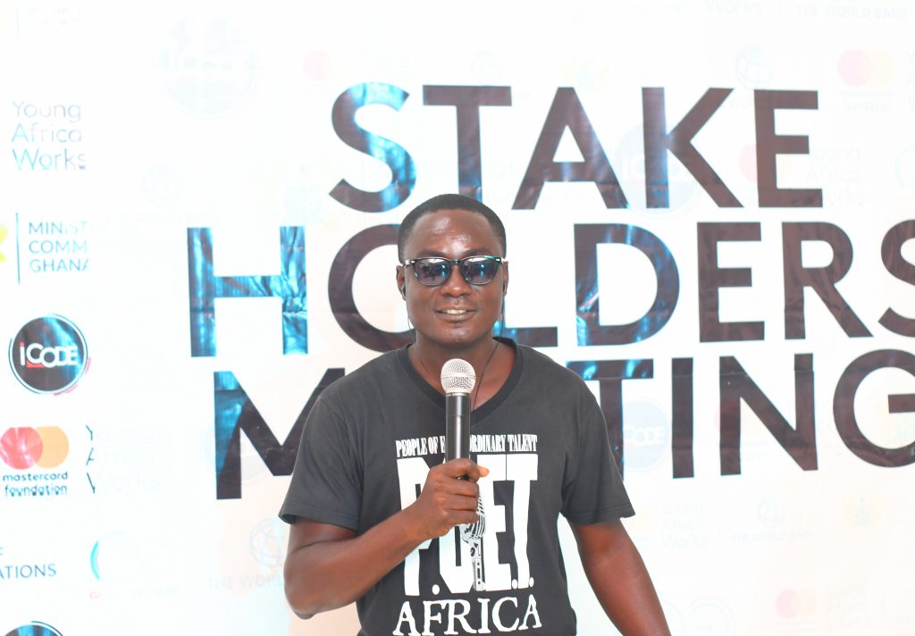 Young Black man with dark glasses holding a microphone with a tee shirt that reads 'POET' in front of a sign reading 'Stakeholders Meeting.'