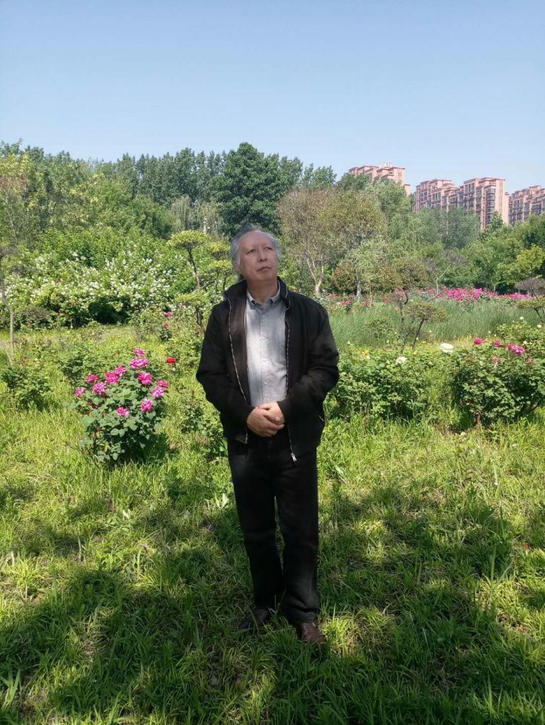 Older Asian man standing in a green field with flowers with large apartment buildings in the background