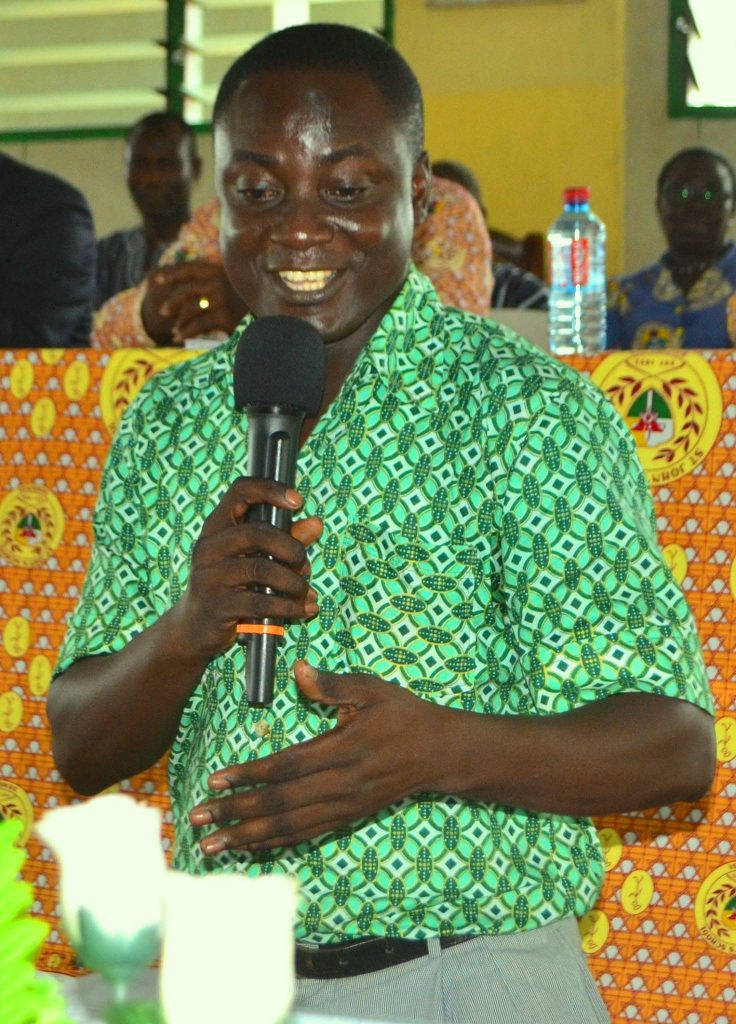 Young black man with a green patterned shirt speaking into a microphone in a gathering of people indoors. 
