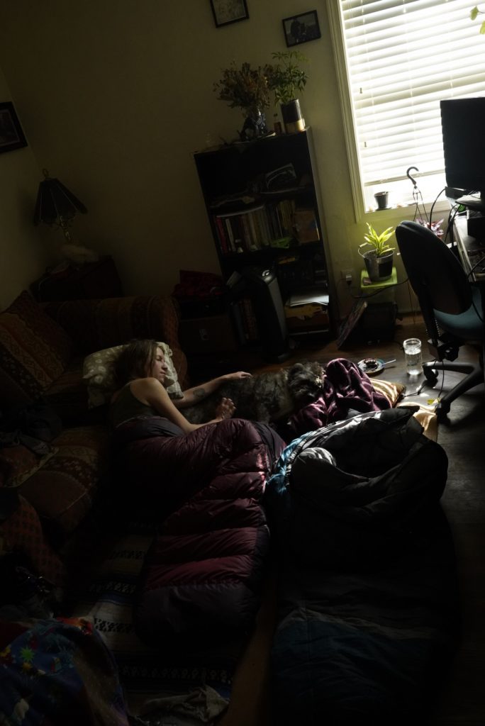 Woman resting on a couch in a dark room with a blanket over her and the curtains closed. Light from a window illuminates a green houseplant. 