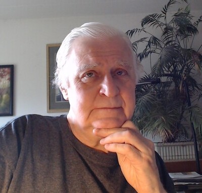 Older white man posing in his home near a houseplant and some paintings on the wall. 
