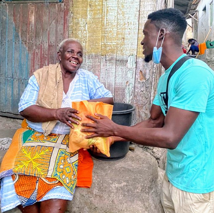 Young man, standing, handing a bag of food to an older woman seated outside on the porch of a building. She's got a colorful yellow and orange skirt. 
