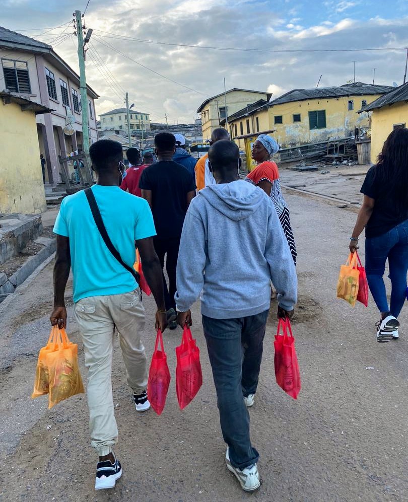 Group of young people carrying bags of food on a partly cloudy day down a street by some multistory buildings.