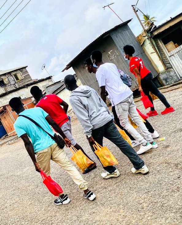 Young people in Ghana in tee shirts, sweaters and jeans and tennis shoes carrying bags of food and walking past buildings. 