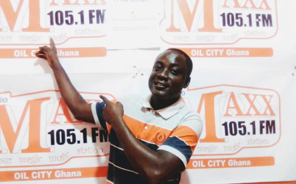 Young Black man in a striped and collared shirt stands in front of a banner for 105.1 FM Radio. His outfit, black and white and orange, matches the color of the sign.