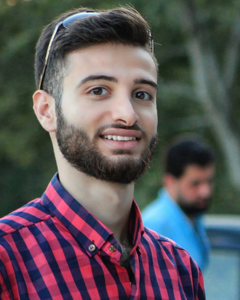 Young Syrian guy, short beard, blue and red checkered polo shirt, sunglasses atop his head of brown hair