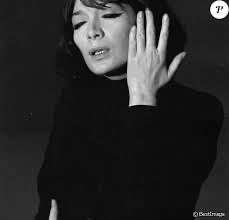 Juliette Greco singing, her eyes are half closed, she's dressed in black and her left hand is up facing her head. 