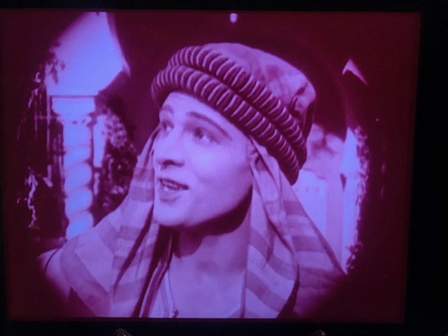 Young white man with a theatrical turban looks off to the left, facing the audience in an old black and white film still image. 