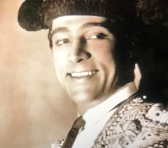 Smiling young-ish white man in a large black hat and a colorful suit, with perfectly white teeth, looking off to the right but facing the audience.