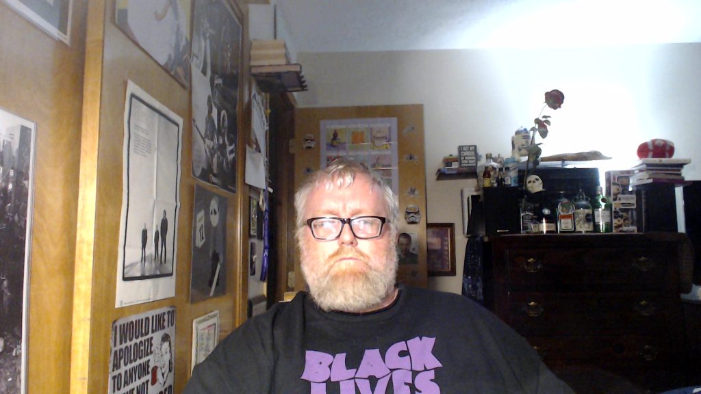 White man with a beard and glasses and a beard and a mustache. He's in a room with some music and movie posters on the walls. He has a Black Lives Matter tee shirt with purple text on a black background.