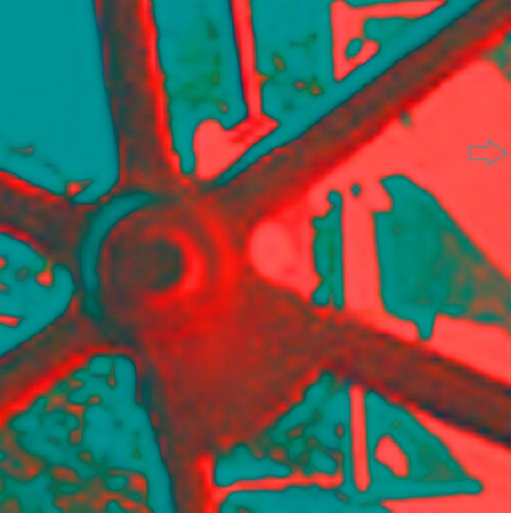 Image of a red train wheel up close, part of the wheel with the spoke off center to the left as the central focus point of the image. Background is light blue and the photo is hazy like an underdeveloped negative. 