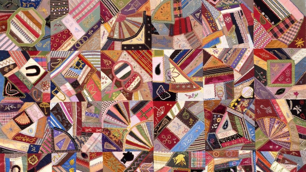 Quilt image, various kinds of fabric joined together at varying angles in a grid of squares. Pink, brown, light and dark blues, gold and yellow and pink. 