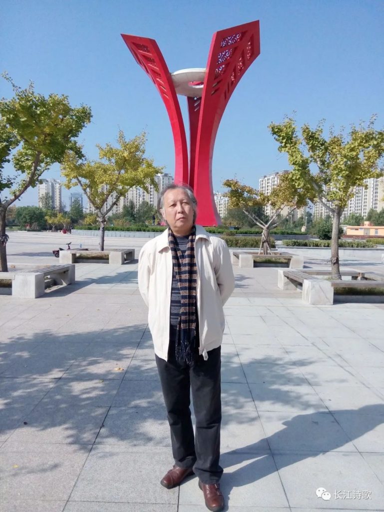 Middle aged Chinese man in a tan jacket and black pants and a scarf standing on a city sidewalk in front of some trees and a tall red sculpture