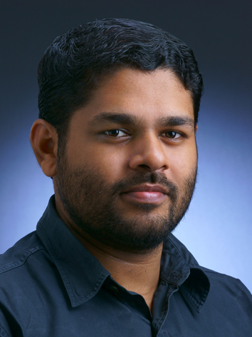 South Asian middle aged man with brown hair and a small beard. Blue collared shirt.
