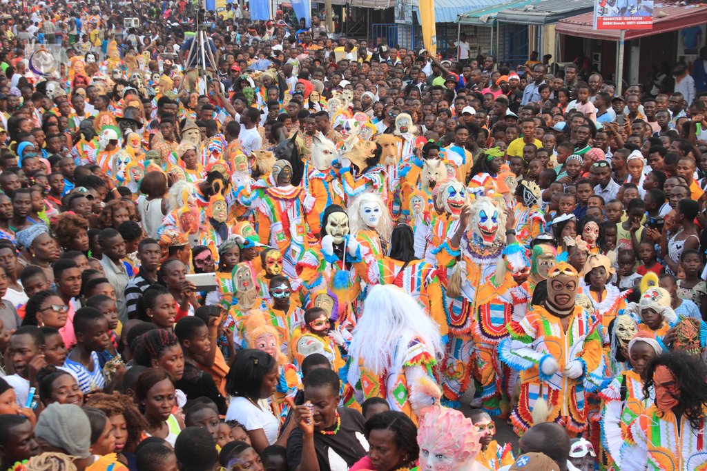 Large masquerade parade in Ghana with the people in costumes and full-face masks. Spectators in tee shirts crowd on either side to watch. 