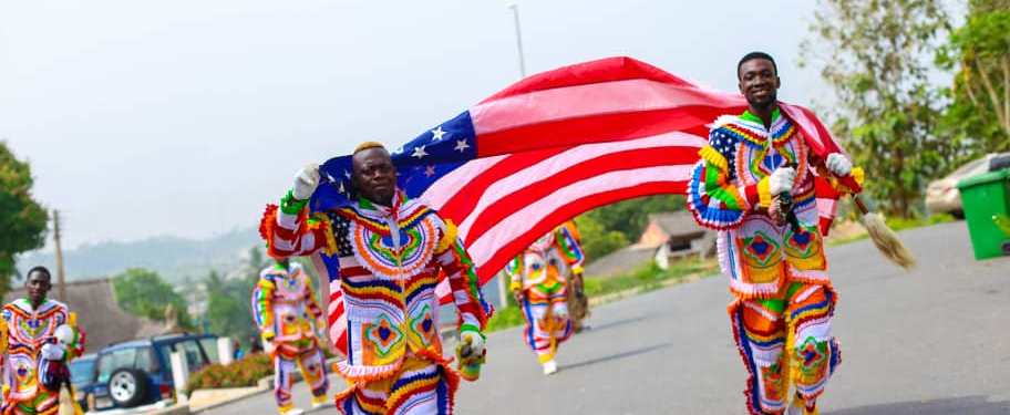 Two young Black men in the foreground carrying a large red, white and blue American flag, dressed in multicolored clothing. Others in similar getup are behind them, on the road in a suburban street. 