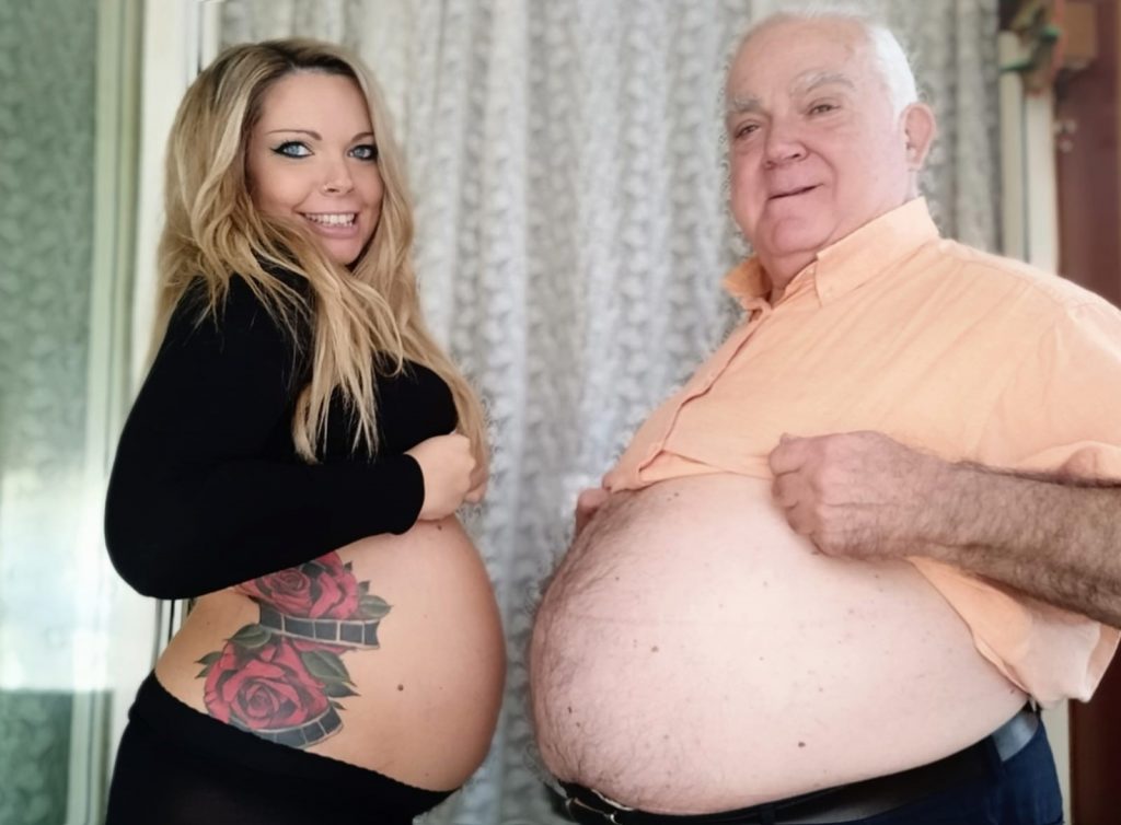 Older white man with thinning hair, a light orange collared shirt and a large chubby belly, uncovered. Next to him is a young white woman, with a black top and black pants. She has also uncovered her pregnant belly and has a tattoo on her side of red roses and what looks like film reels. 