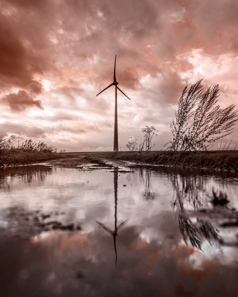 Single windmill in a wet marsh area with water and some windswept grassy plants, clouds overhead and a pink/purple sunset or sunrise color.