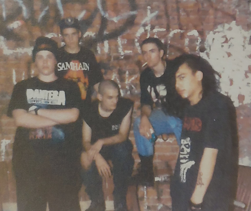 Five guys, some white and some of indeterminate race, with black tee shirts with heavy metal band logos, posing in front of a brick wall with graffiti.