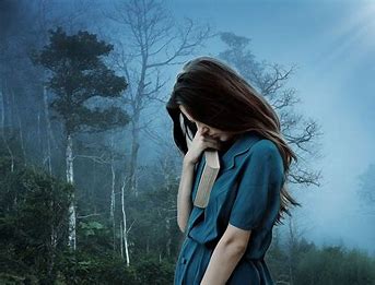 White woman with dark hair and a blue dress bent over in thought, with a book under her arm. She's in a misty forest with some trees which have leaves and others which don't. 