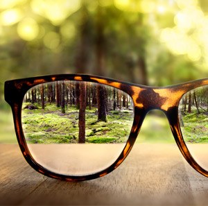 Fuzzy image of far-off trees made brighter and clear through the lens of glasses on a wooden table. 