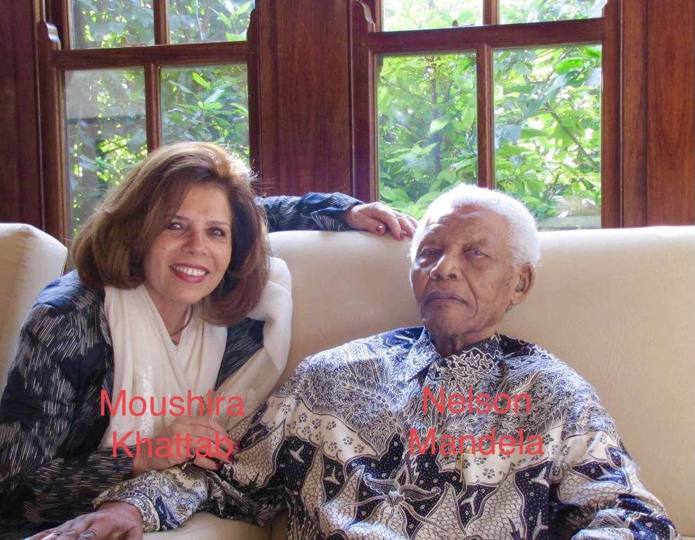 Light-skinned middle aged woman with brown shoulder-length hair sits on a white couch in a living room with windows and green plants outside. She's wearing a white scarf and patterned blue and white top. Nelson Mandela, a white haired Black man, sits next to her and is in a blue and black and white patterned collared shirt. 
