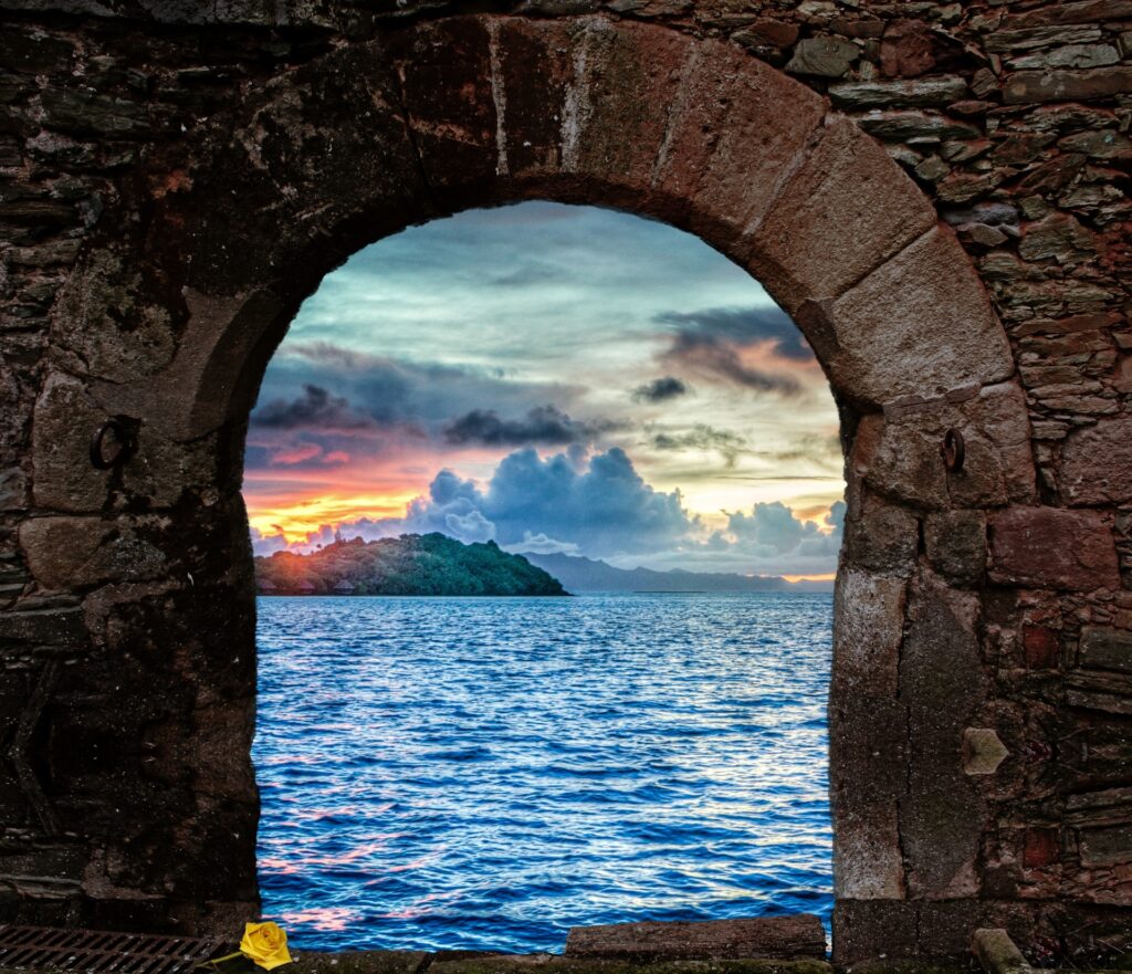 Arched opening in a brick wall opens to a view of a large body of water with clouds and a sunrise/sunset in the background. A green island looms in the distance. 
