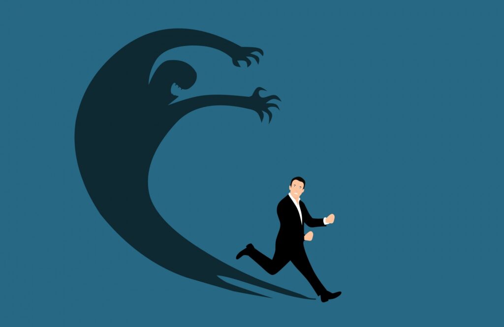 Nondescript clip art white male figure in a business suit runs from his shadow, which has grown and morphed into a menacing creature with teeth and claws. 