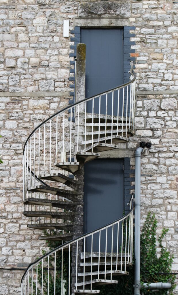 Brick building with white stone bricks and a gray painted door. A spiral concrete staircase with a railing extends out of the door as a fire escape. 