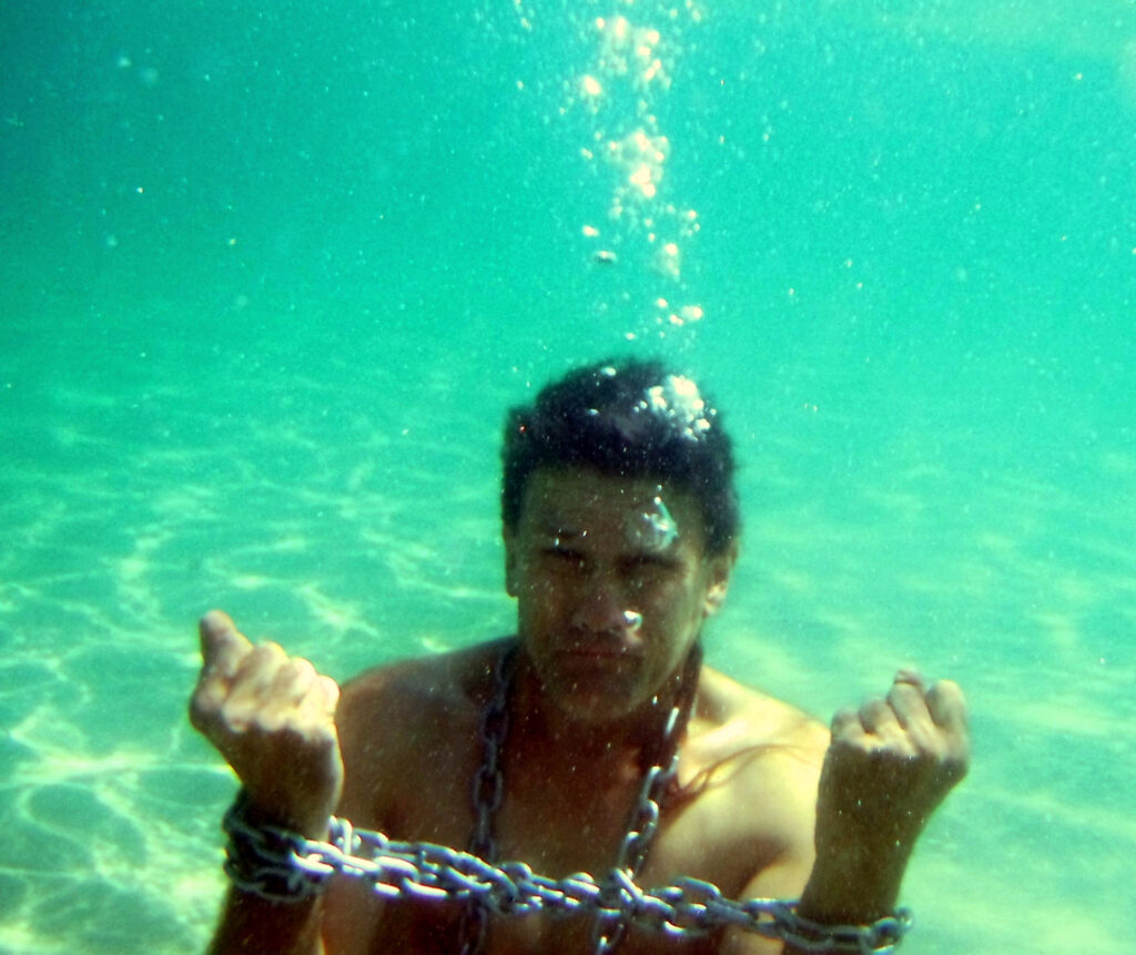 White or Latino man with short brown hair (real image of a person) with chains around his neck and hands underwater. Part of an escape stunt game. 