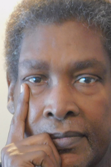 Middle aged Black man with short hair and brown eyes. He's got a hand on his chin and is facing the camera.