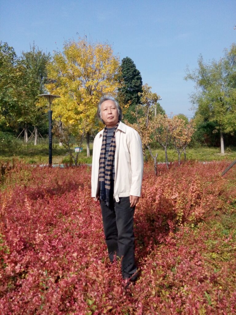 Older Chinese man with a white coat and scarf and black jeans standing in a field with a reddish-brown leaved shrub behind him and a yellow tree and some green trees and a lamp behind him. 