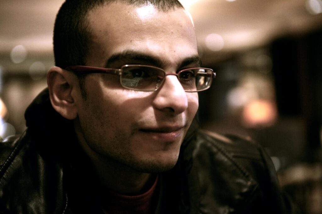 Egyptian man with glasses and a black or brown jacket in half-light. 