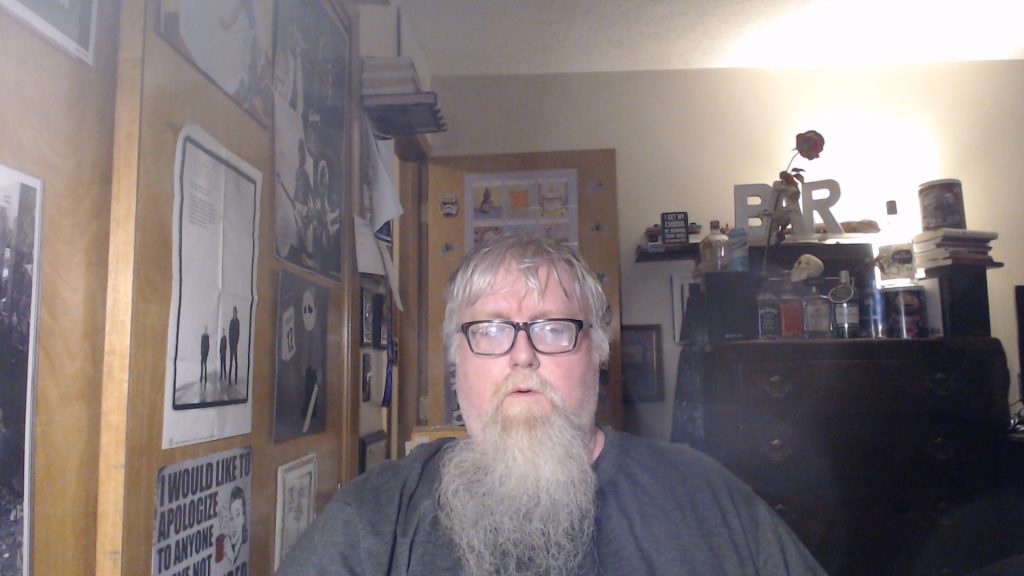 Middle aged white man with glasses and a beard stands in a room in front of speakers and movie and band posters. 