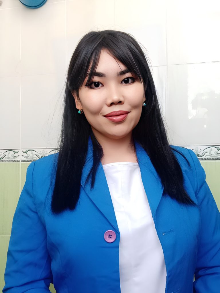 Young Central Asian woman with brown eyes and long straight black hair. She's got earrings and makeup on and is wearing a white top and a blue jacket.