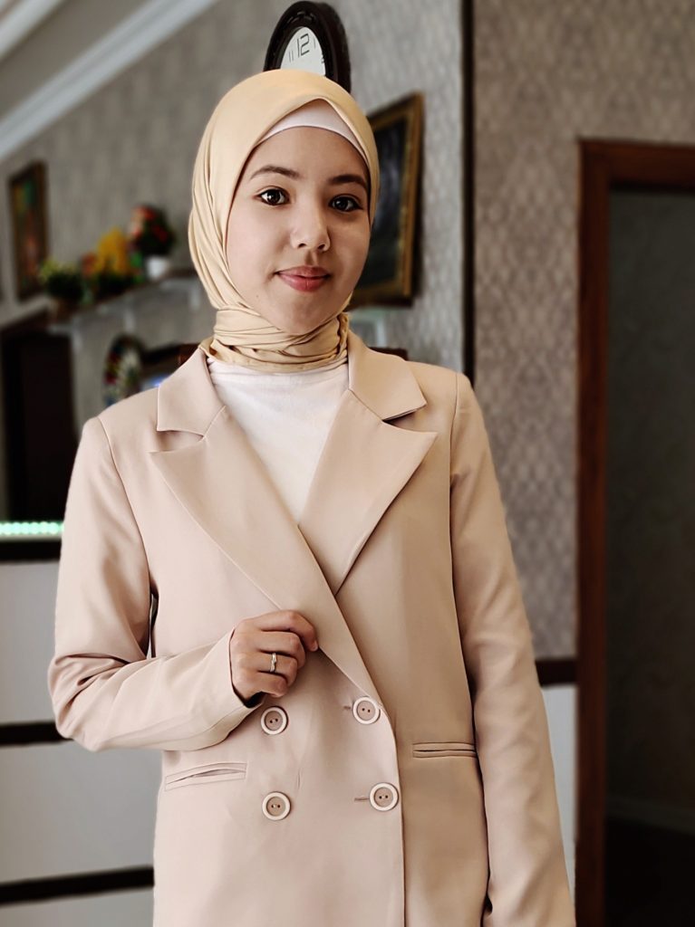 Young Central Asian woman with a peach headscarf and coat over a cream colored top. She's standing in a living room with pictures on the wall and a clock behind her. 